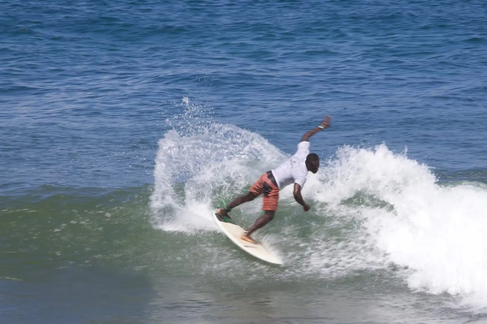 A surfer in a white t-shirt and orange shorts leans into a sharp turn on a green wave, throwing up white spray.