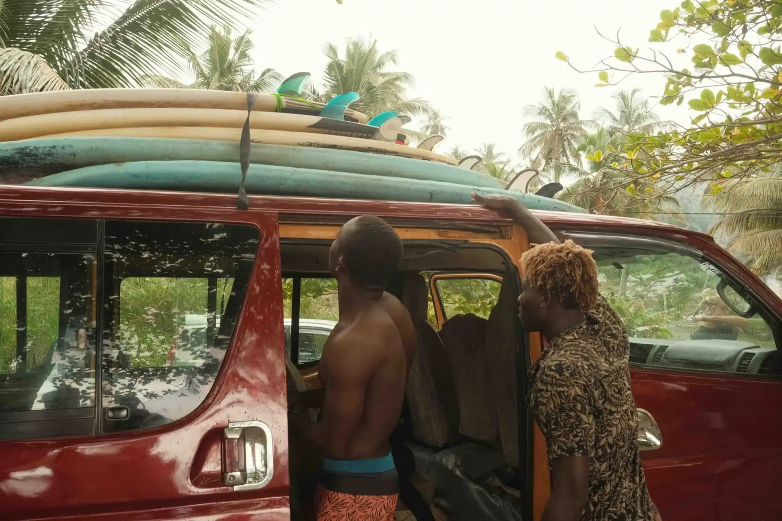 Two people look up at surfboards stacked on the roof of a red minibus parked among palm trees.