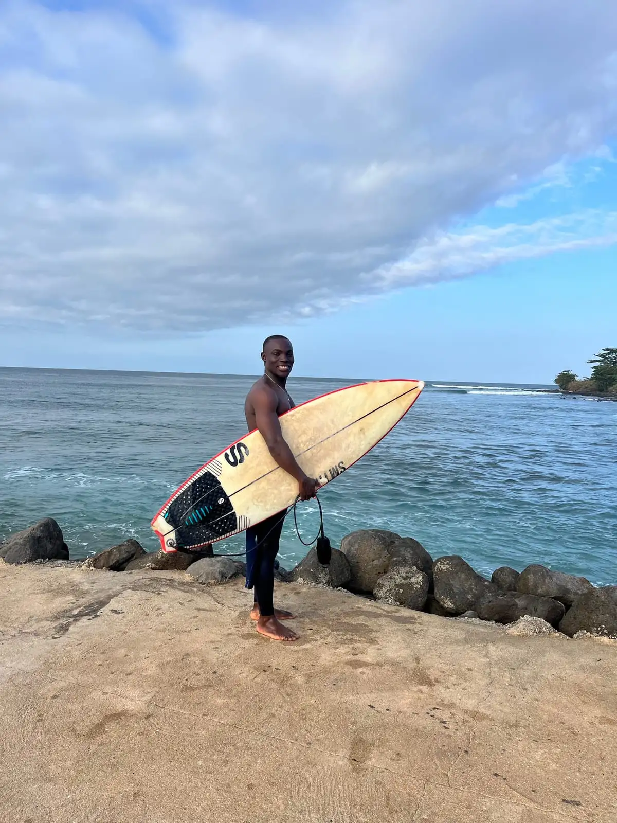 A smiling surfer stands on a rocky point holding a surfboard, with blue sea and a cloudy sky behind.