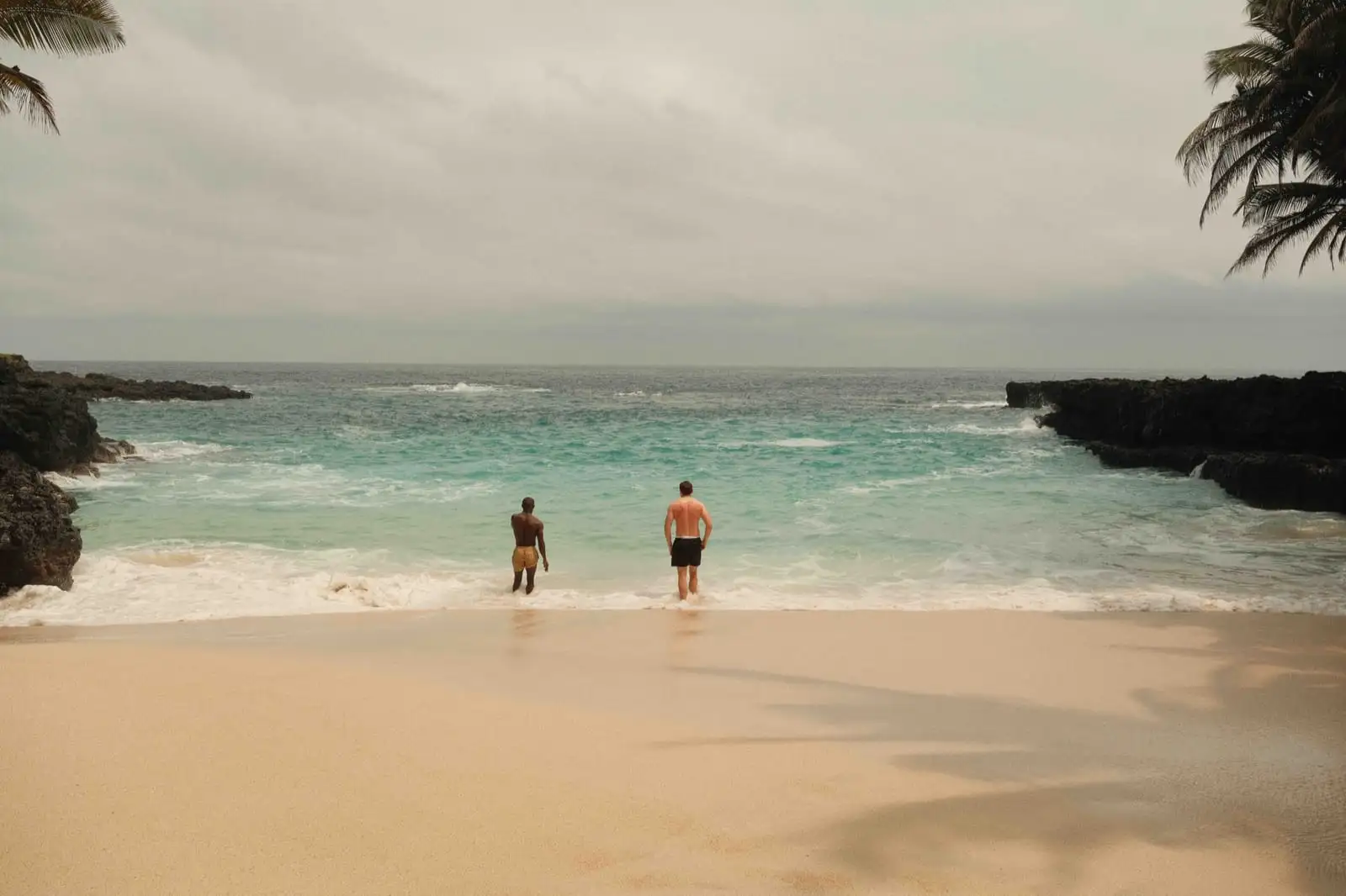 Two people wade into turquoise water at the edge of a sandy beach between dark rocks.