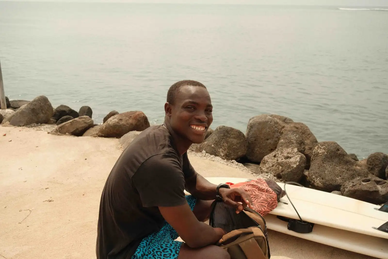 A smiling man sits by the sea near dark rocks, with a surfboard and a backpack beside him.