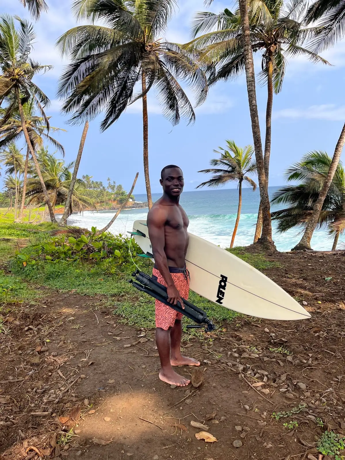 A surfer in patterned red shorts stands on a grassy clifftop holding a surfboard, with palm trees and the sea behind.