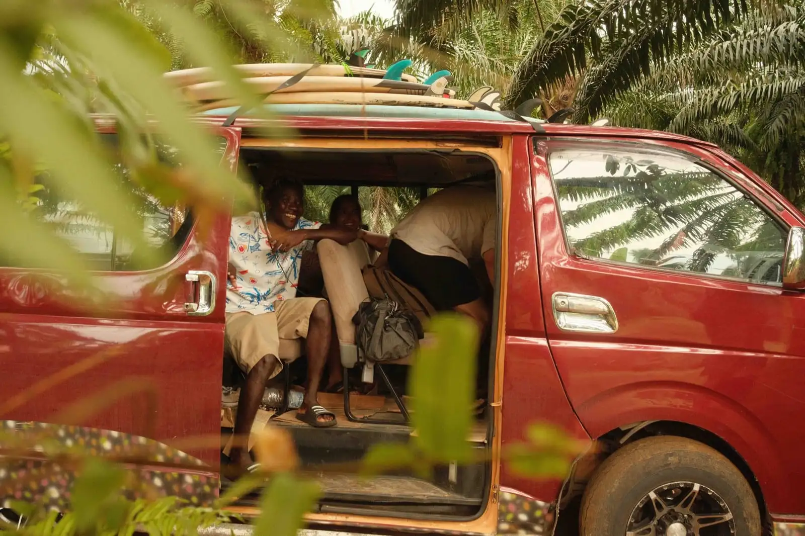People sit inside a red minibus with its side door open, surfboards on the roof and palm leaves in the foreground.
