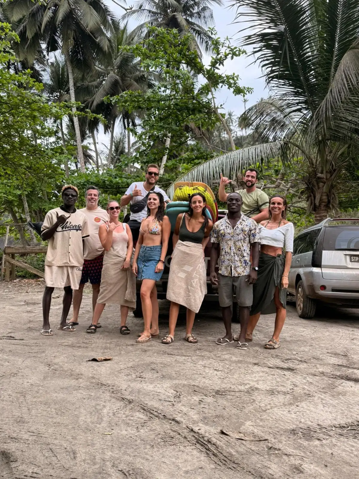 A group of smiling people pose together on a sandy clearing beside a car, with palm trees behind them.