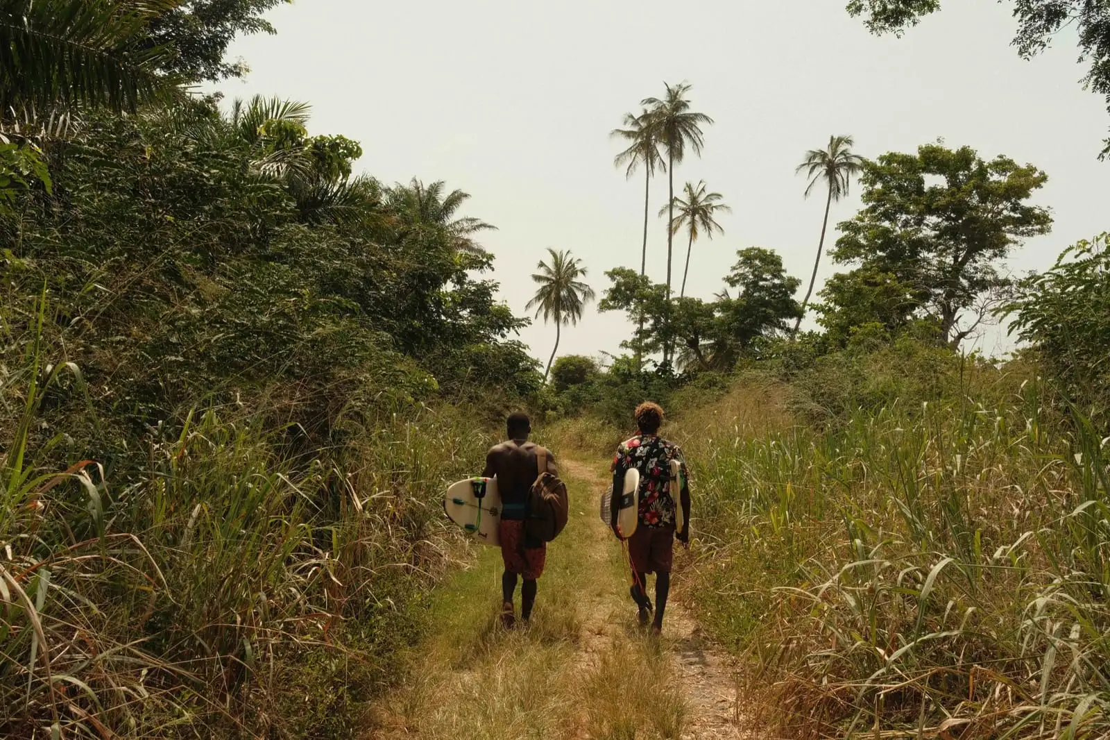 Two people carrying surfboards walk along a grassy track between trees, with palm trees ahead.